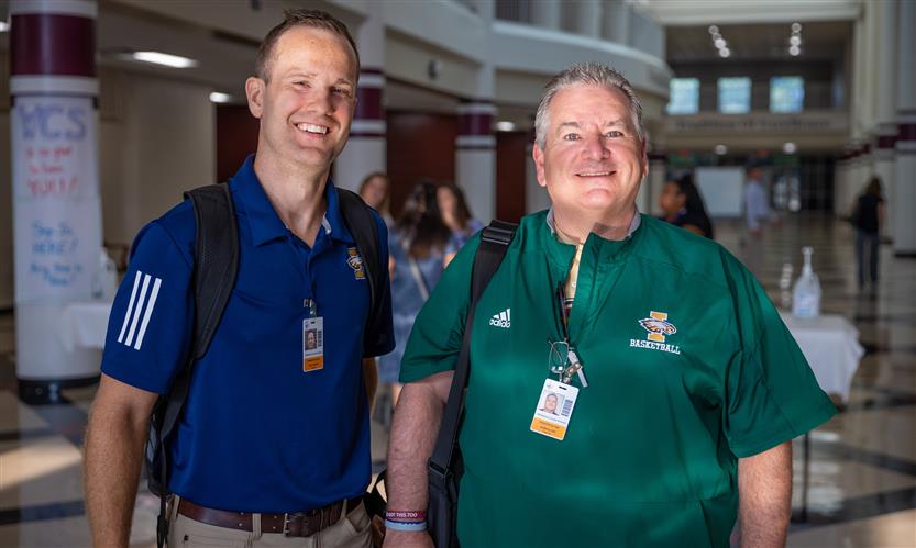 Two men, one wearing dark blue and the other green, smile while standing in the hallway of a school